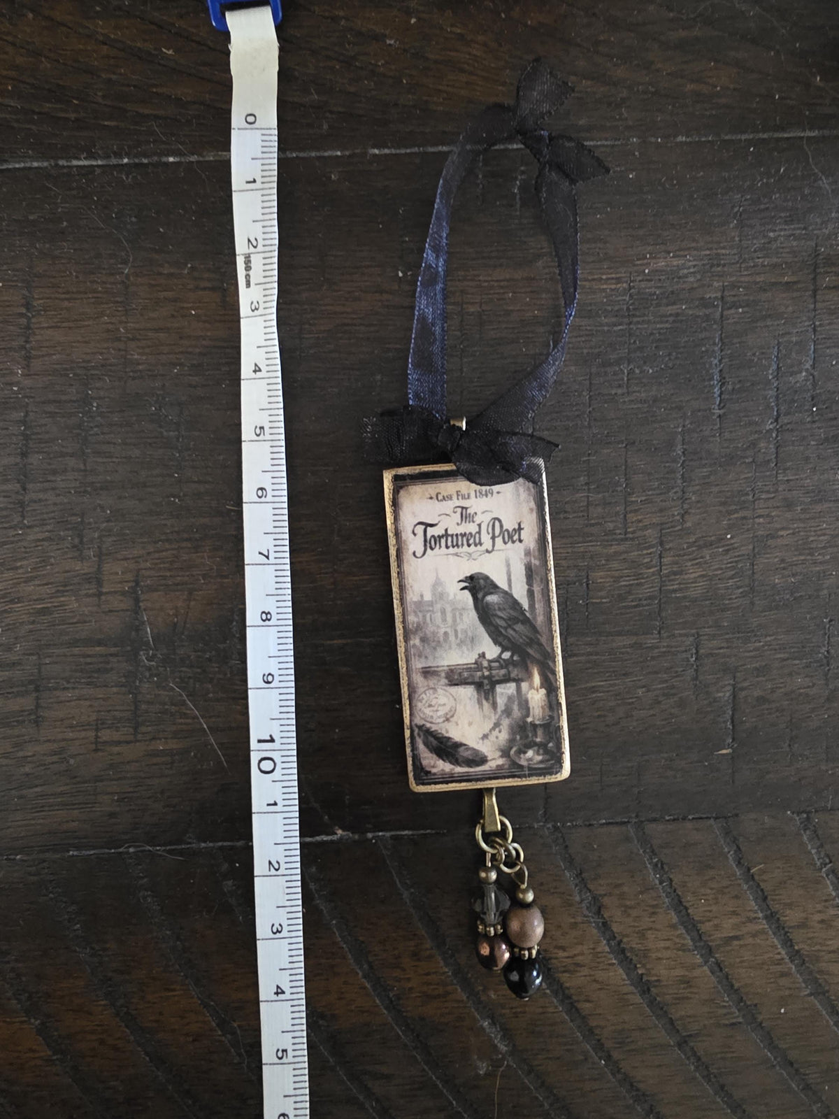 a small, rectangular ornament with a black and white illustration of a bird perched on a branch, hanging from a ribbon. The ornament is placed next to a ruler on a wooden surface.