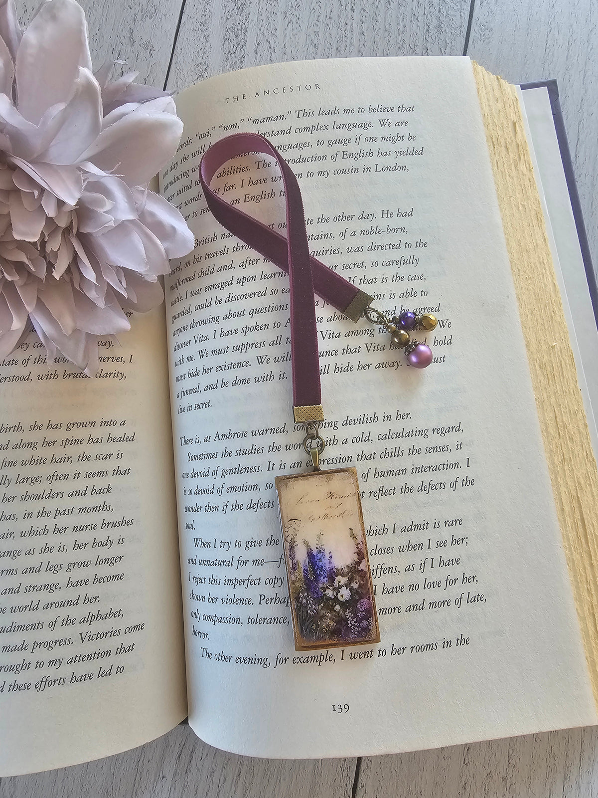 An open book with a purple bookmark featuring a floral design rests on a wooden surface, accompanied by a single white flower.