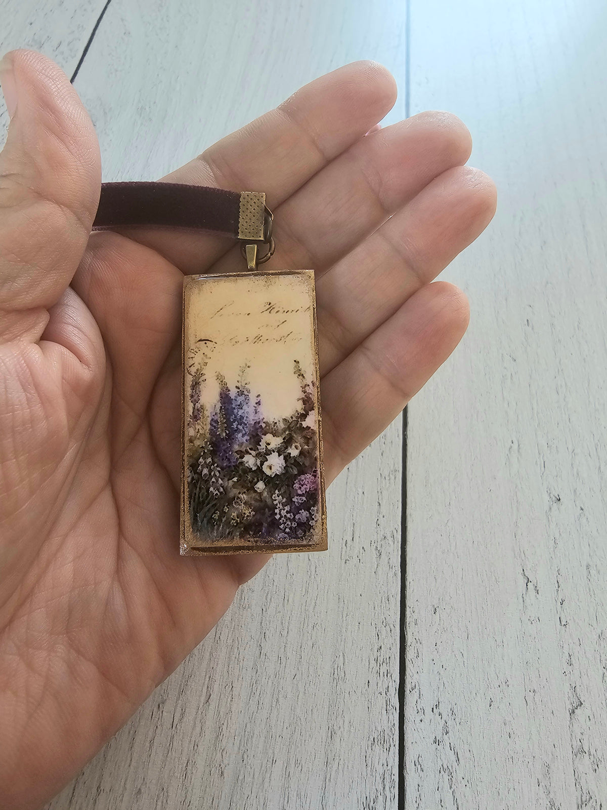 A hand holding a rectangular pendant with a floral design, resting on a wooden surface.