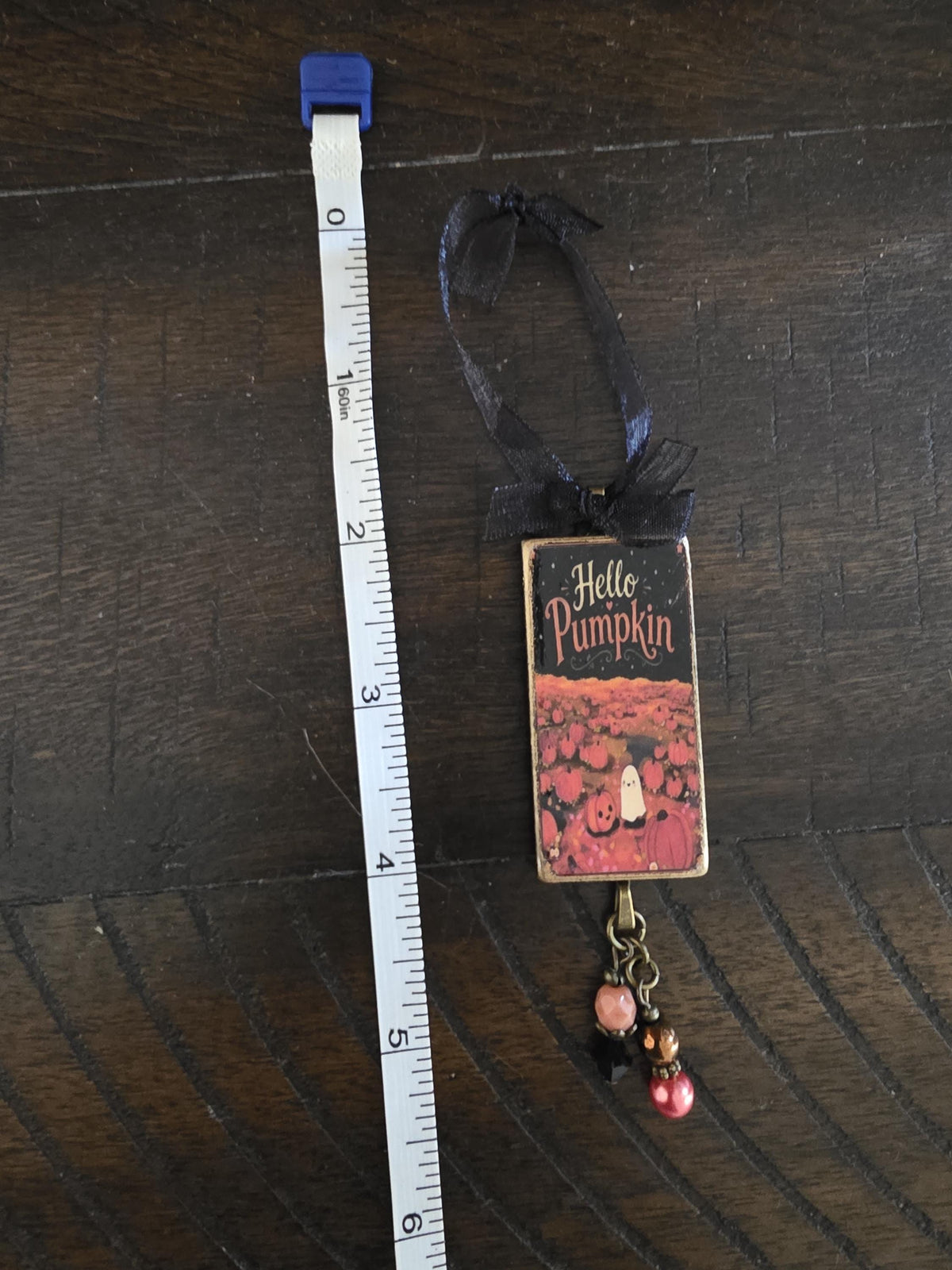 A small decorative item with a black ribbon and a pumpkin-themed design, placed next to a ruler on a wooden surface.
