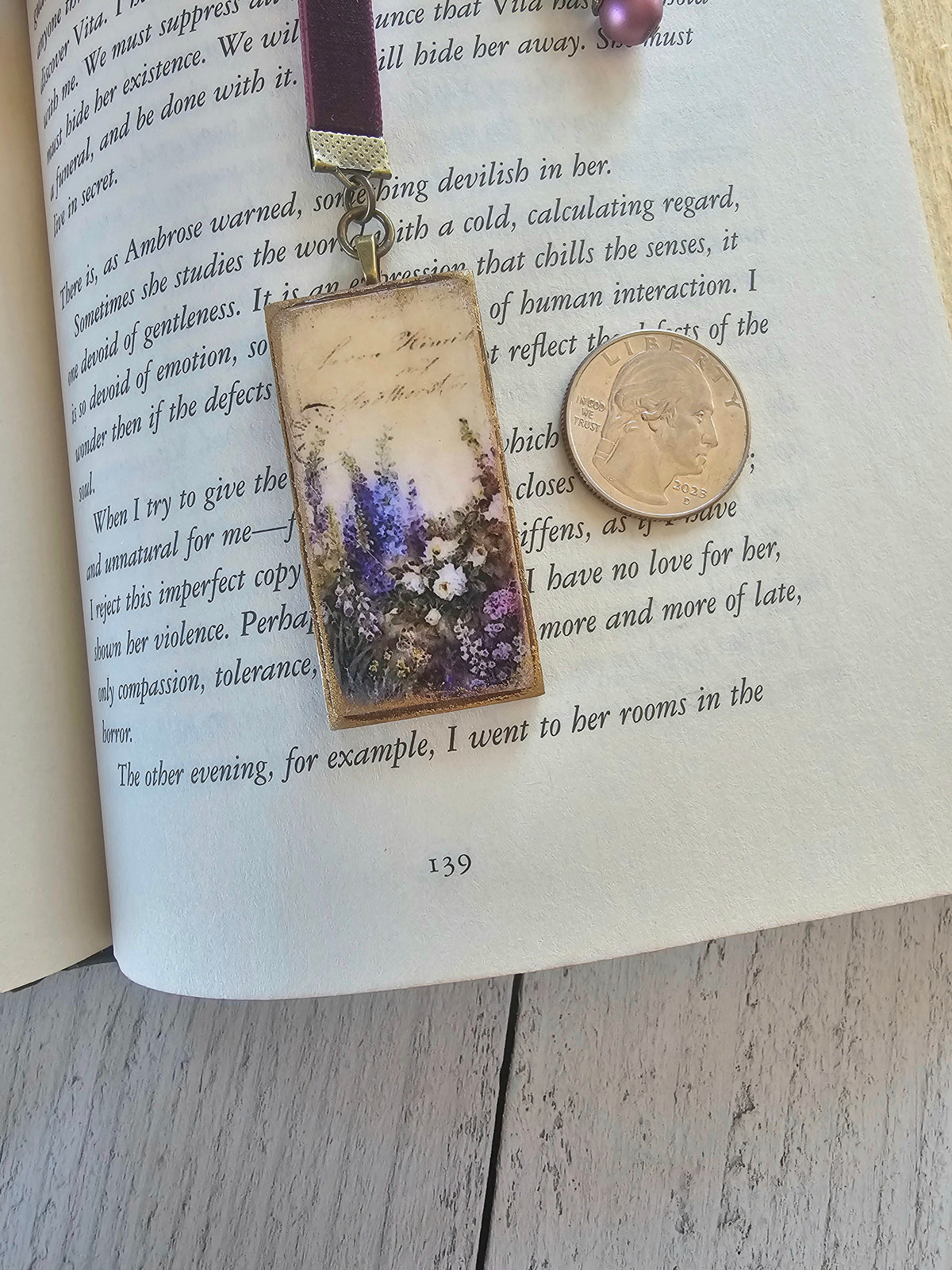 a book with a purple bookmark attached to it, featuring a floral design. The bookmark is placed on top of the book, and there is a coin lying nearby.
