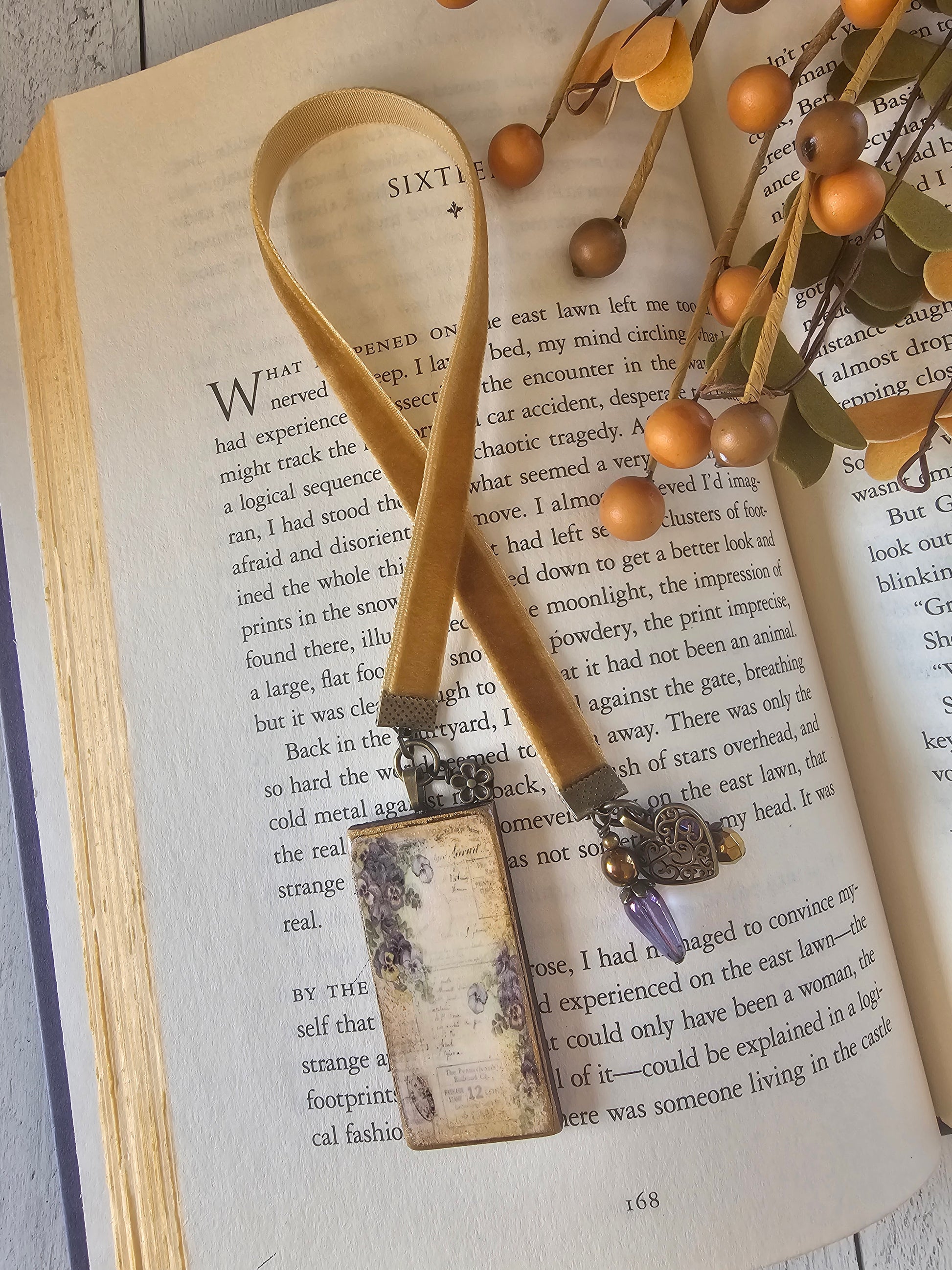 an open book with a beige ribbon bookmark resting on the pages, surrounded by a branch of small orange berries and a small purple charm.