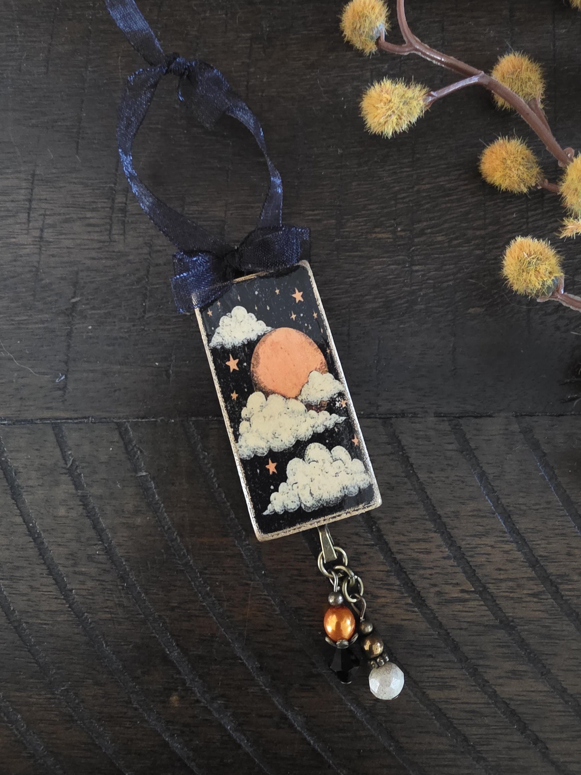 A rectangular pendant with a night sky scene, featuring a moon, clouds, and stars, is suspended from a black ribbon. The pendant is adorned with a small charm and a pearl bead.