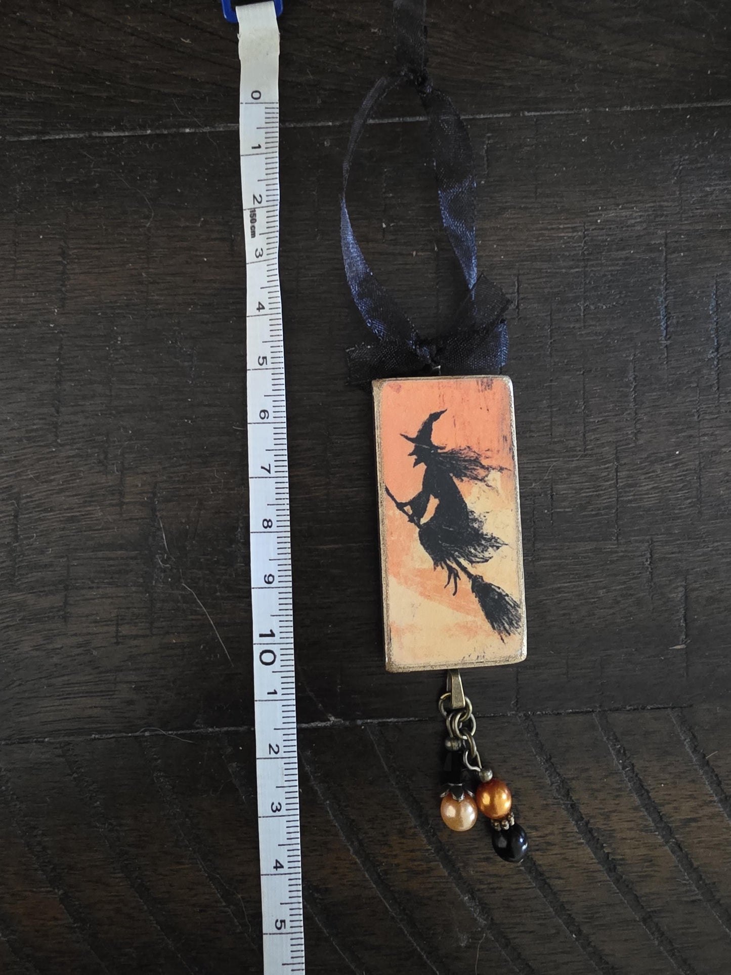 A ruler is placed next to a rectangular charm or ornament that features a silhouette of a witch on a broomstick.