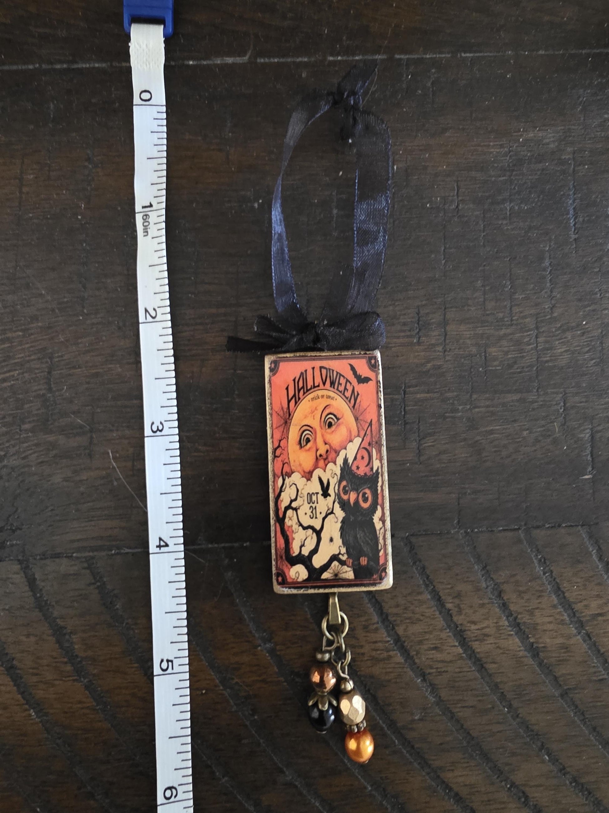 A Halloween-themed ornament with a black ribbon and a small illustration of an owl and a pumpkin. The ornament is hanging from a string with a measuring tape next to it.