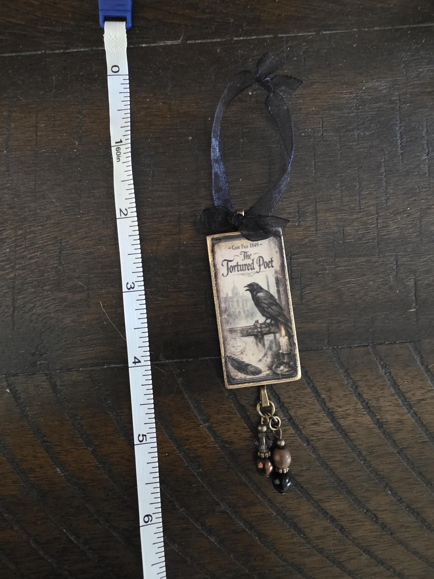 a small, ornate ornament with a bird on it, hanging from a ribbon. The ornament is placed next to a ruler on a wooden surface.