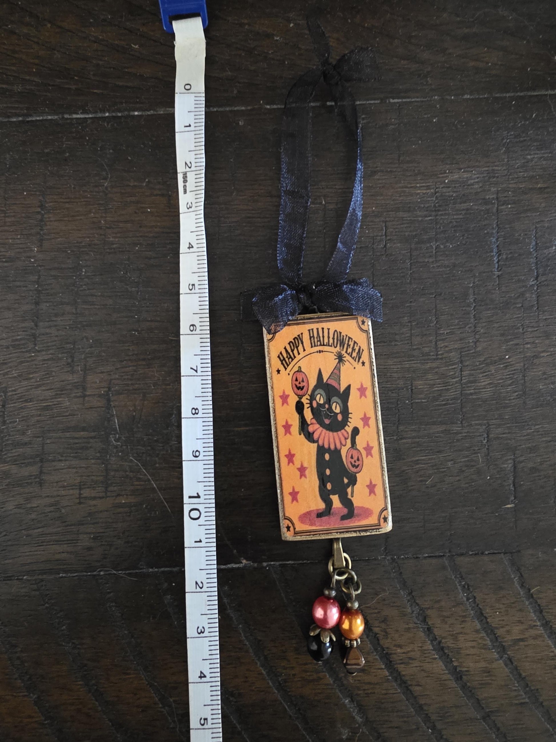 A Halloween-themed ornament featuring a black cat wearing a party hat and holding a pumpkin, with the text "Happy Halloween" displayed on the ornament.