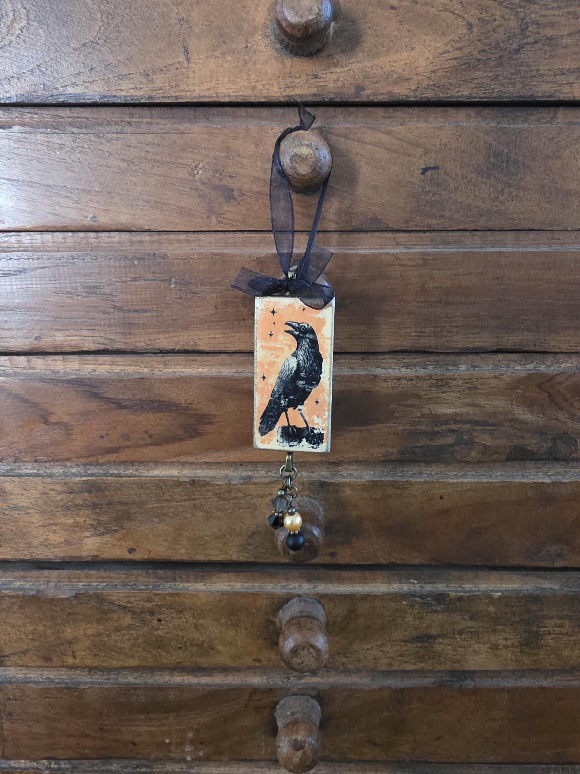 a wooden drawer with a small, rectangular, and ornate piece hanging from it. The piece has a black bird illustration on it, and it is decorated with a black ribbon.