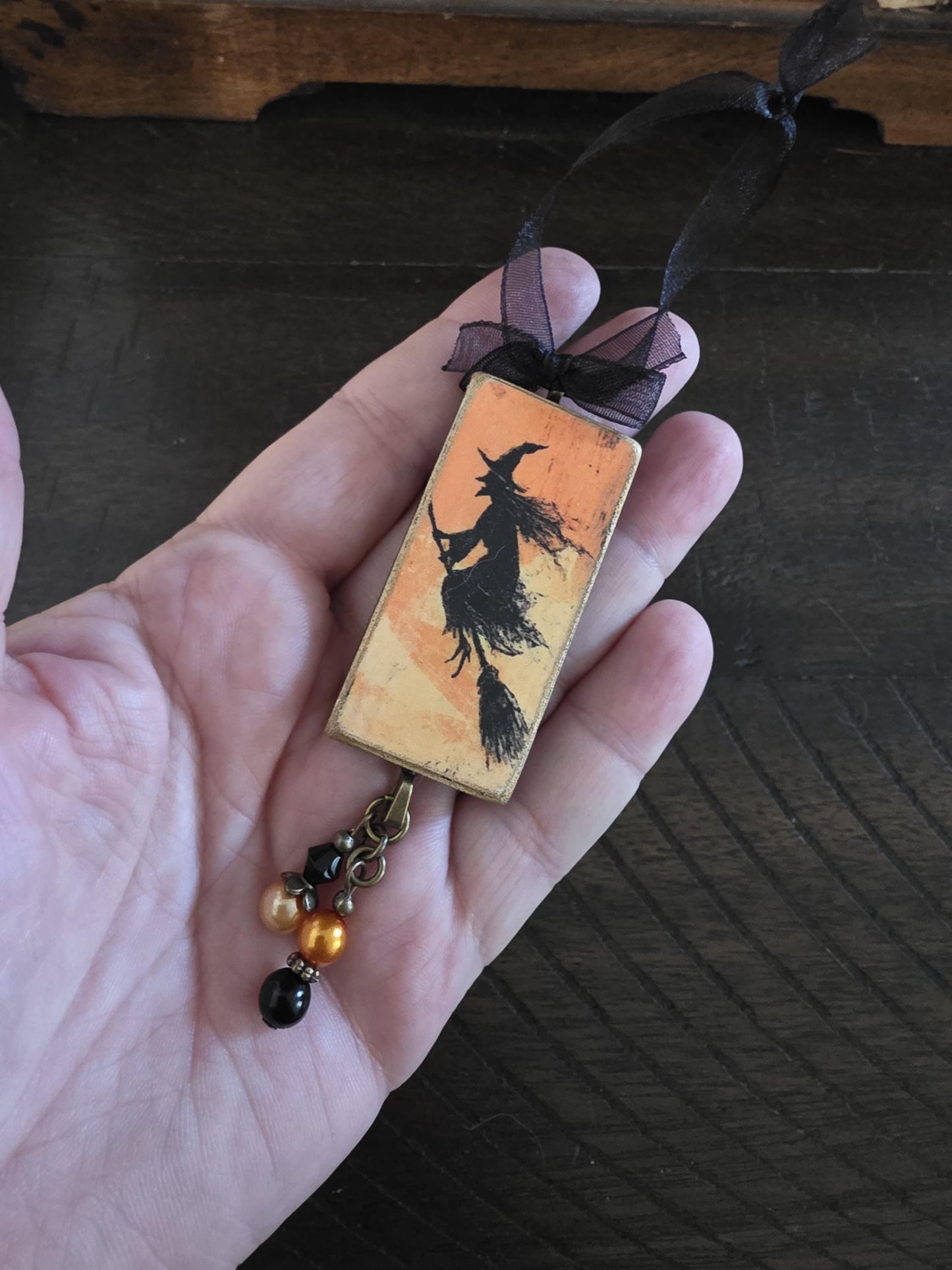 A hand holding a rectangular pendant with a black silhouette of a witch on it, along with a black ribbon and small beads.