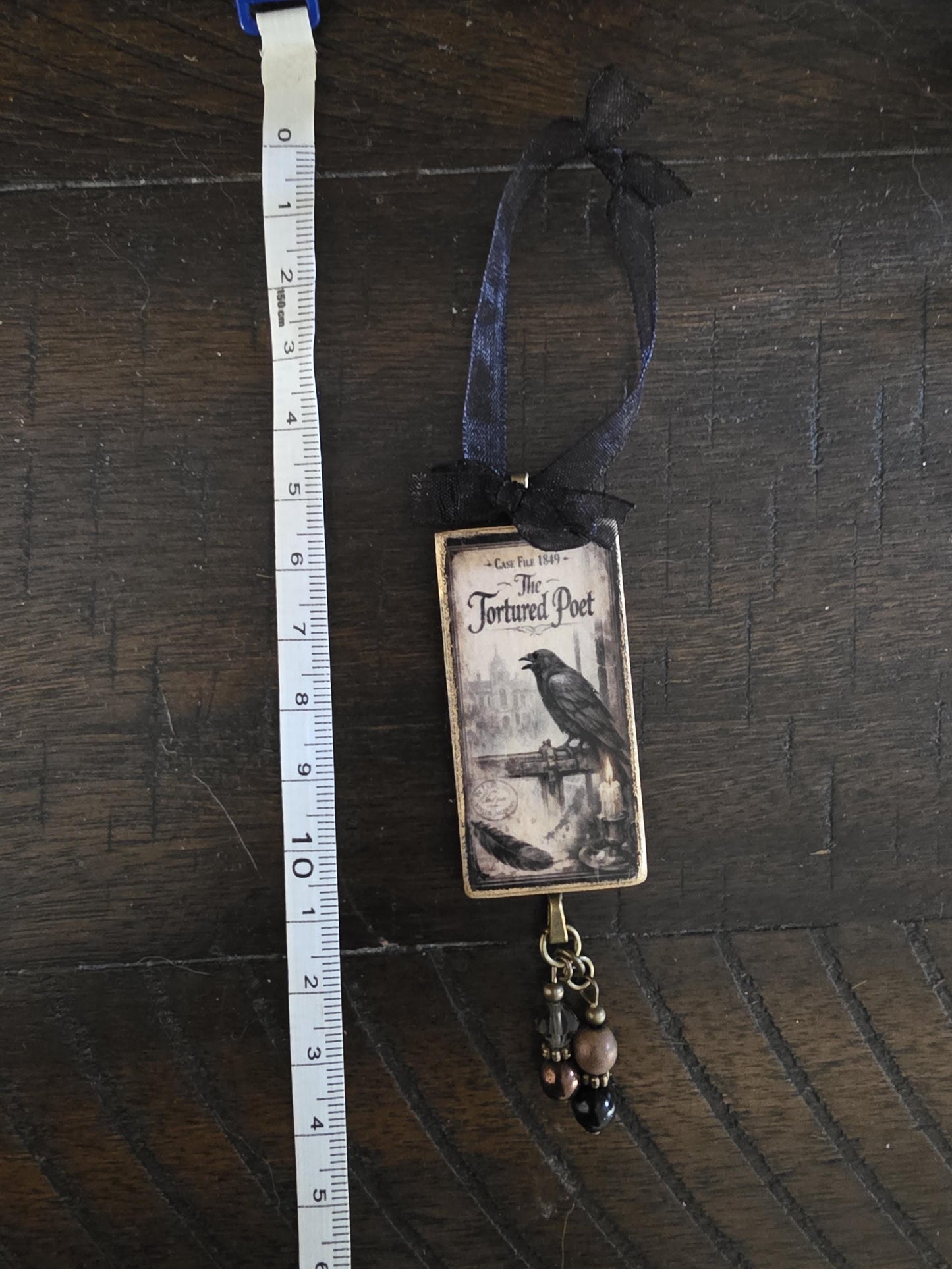 a small, rectangular ornament with a black and white illustration of a bird perched on a branch, hanging from a ribbon. The ornament is placed next to a ruler on a wooden surface.