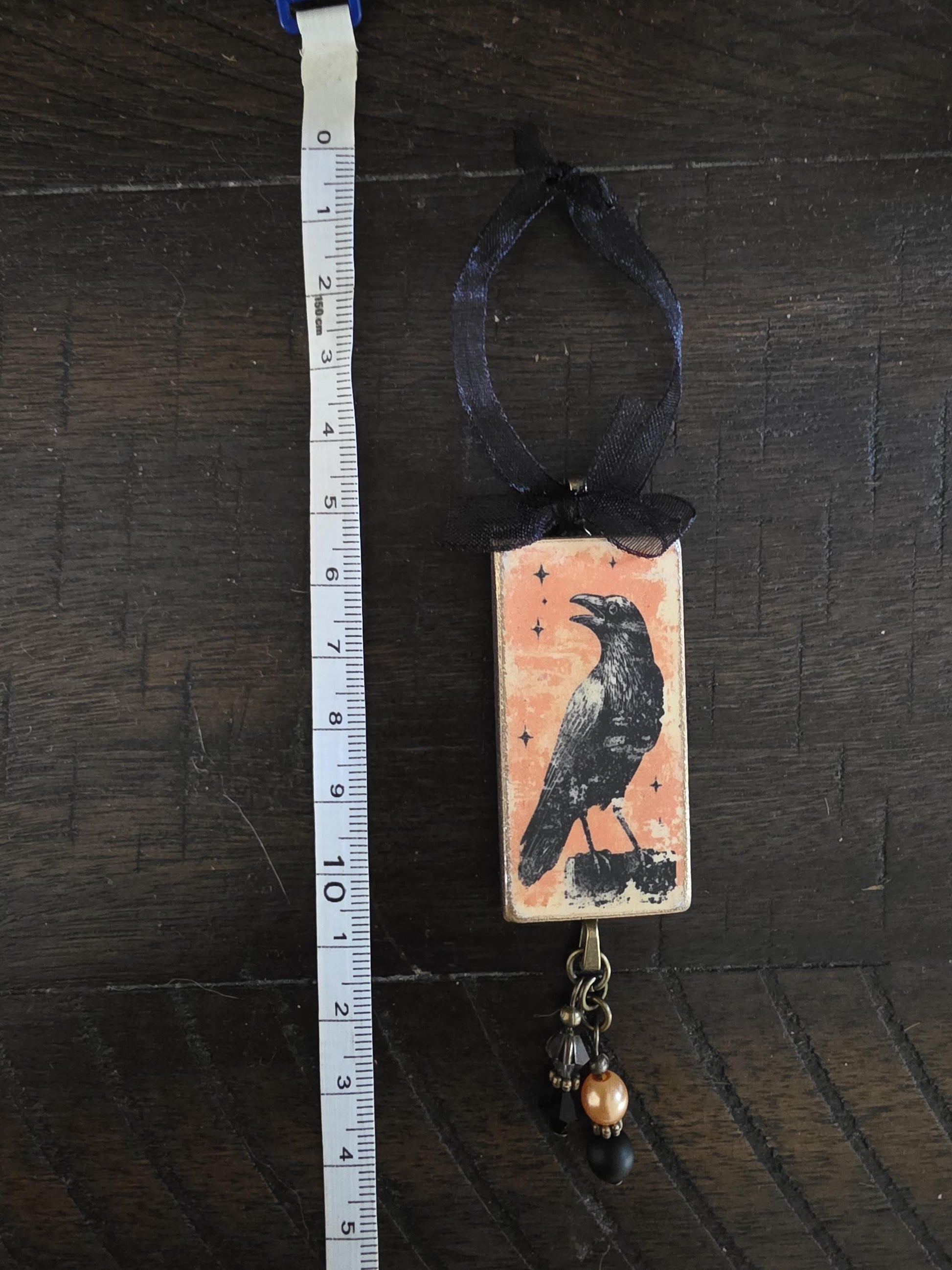A ruler is placed next to a rectangular pendant featuring a black bird illustration.