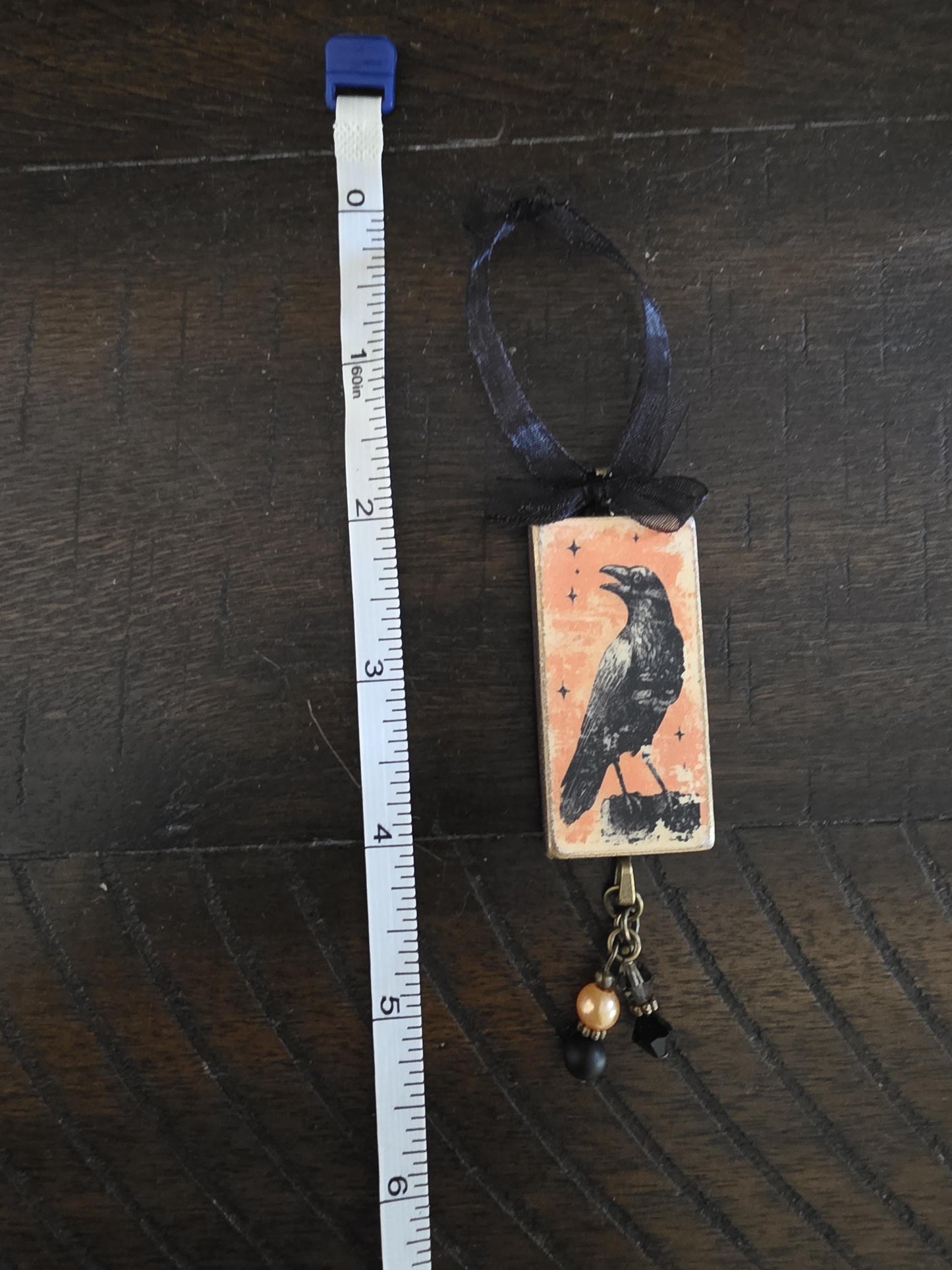 a crow-themed pendant hanging from a ribbon, with a ruler placed next to it on a wooden surface.