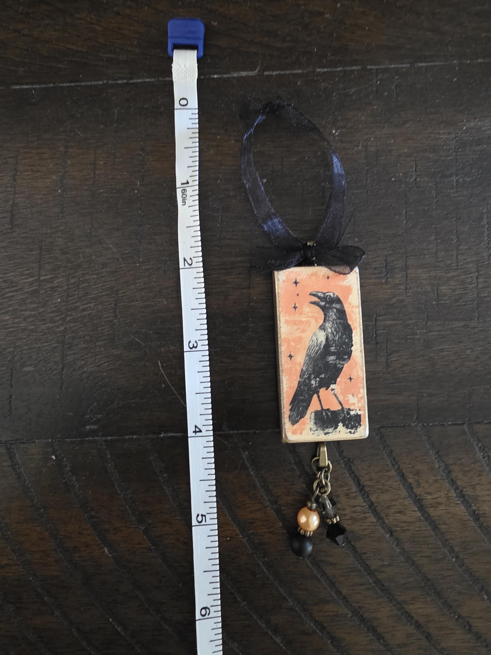 a crow-themed pendant hanging from a ribbon, with a ruler placed next to it on a wooden surface.