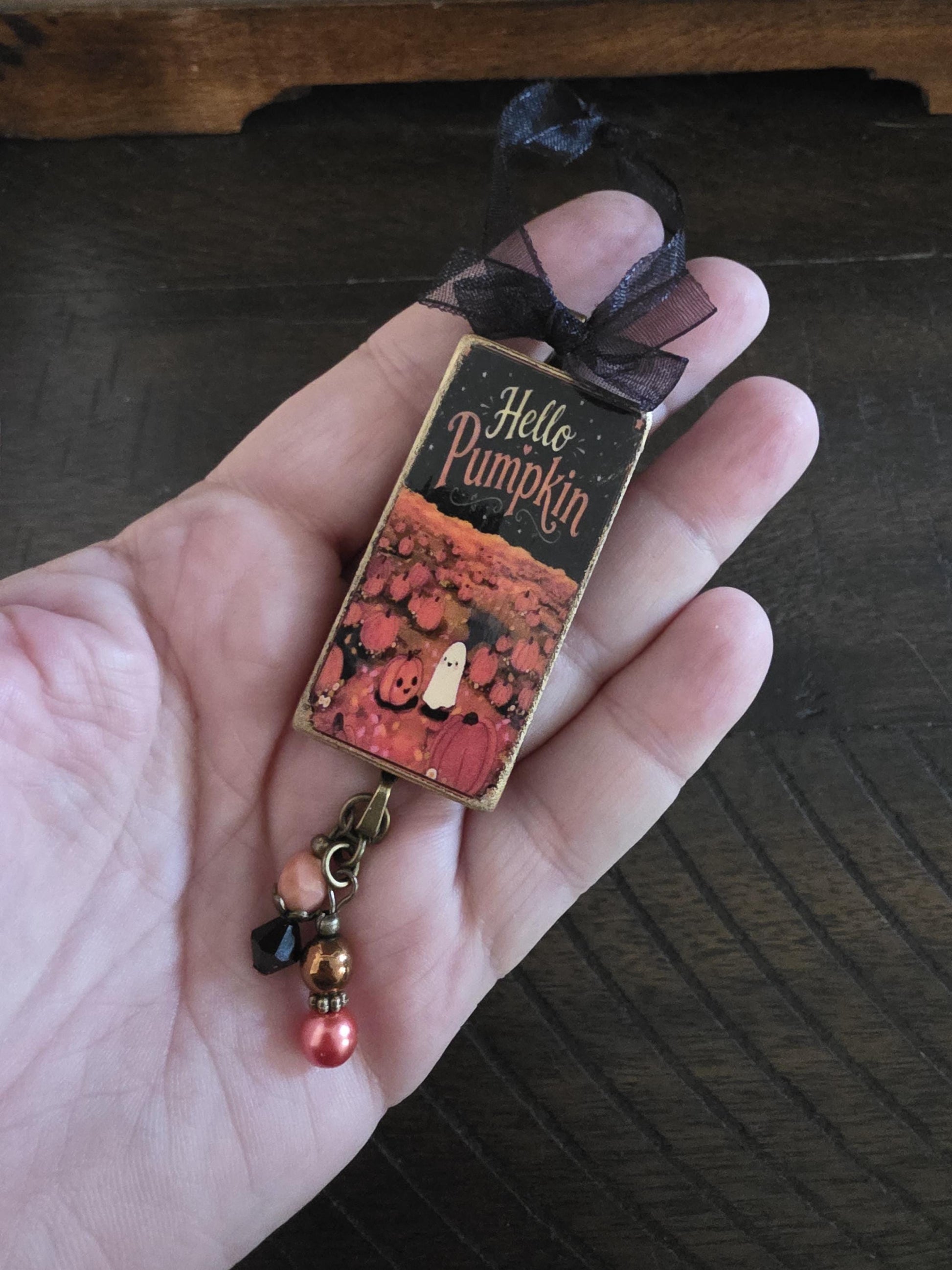A hand holding a small decorative item with a pumpkin and ghost design, and a ribbon around it.
