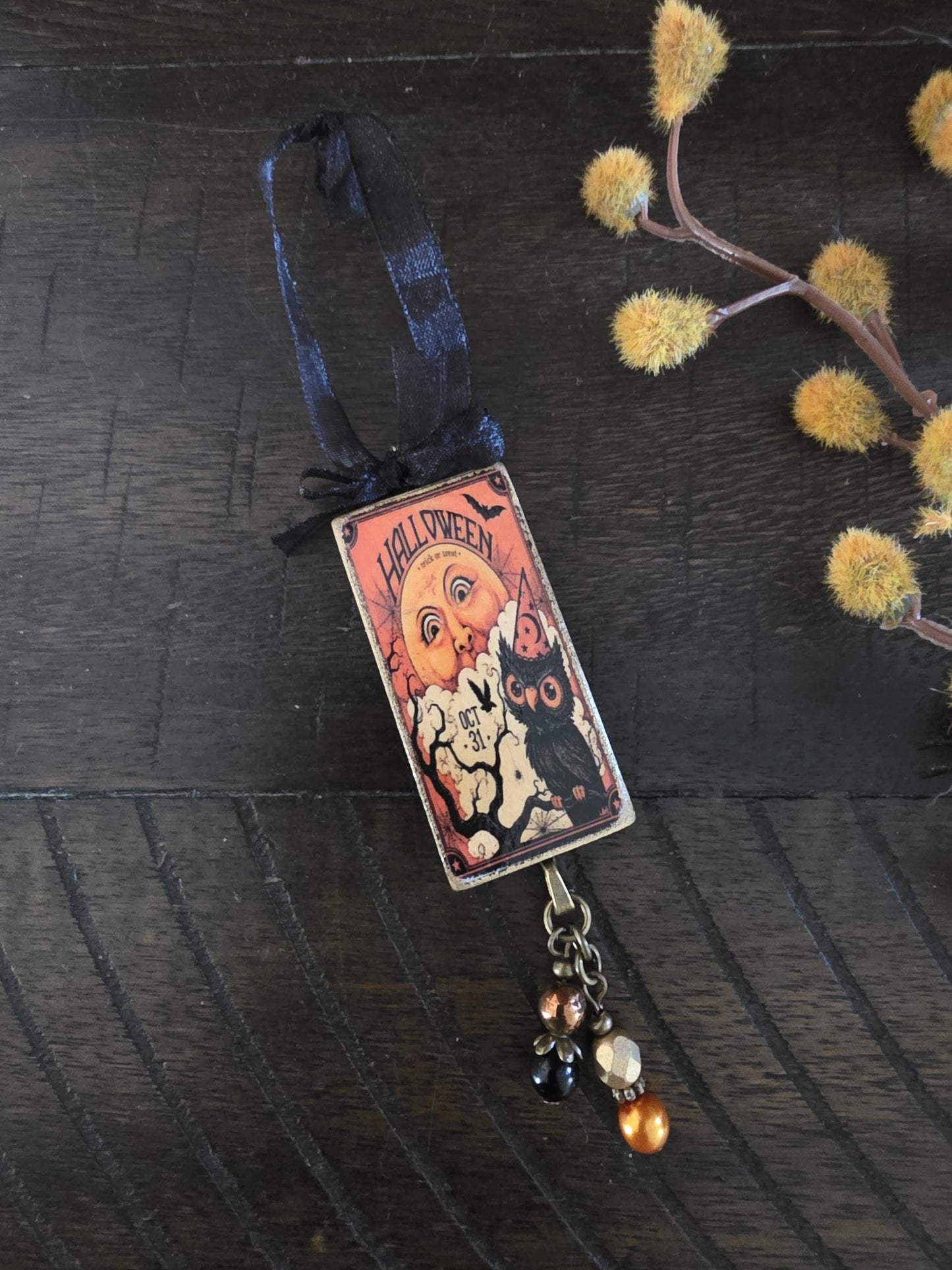 A Halloween-themed ornament with a black ribbon and a small cat illustration is displayed on a wooden surface, accompanied by a branch of yellow flowers.