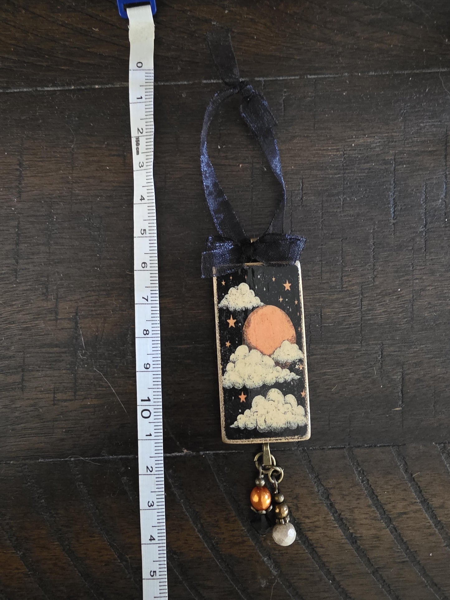 A decorative pendant hanging on a wooden surface, featuring a night sky scene with a moon, clouds, and stars. The pendant is accompanied by a ribbon and has a small charm hanging from it.