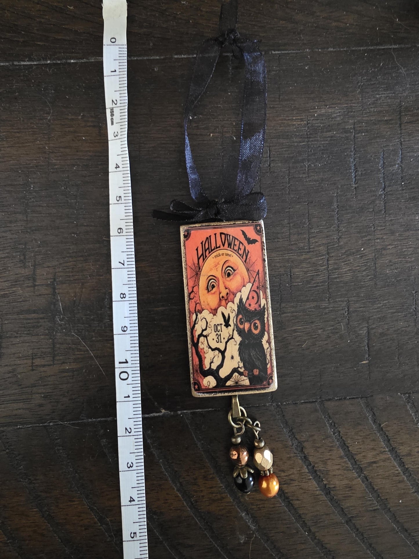 A Halloween-themed pendant hanging from a ribbon, featuring an owl and a bat, is placed next to a ruler on a wooden surface.