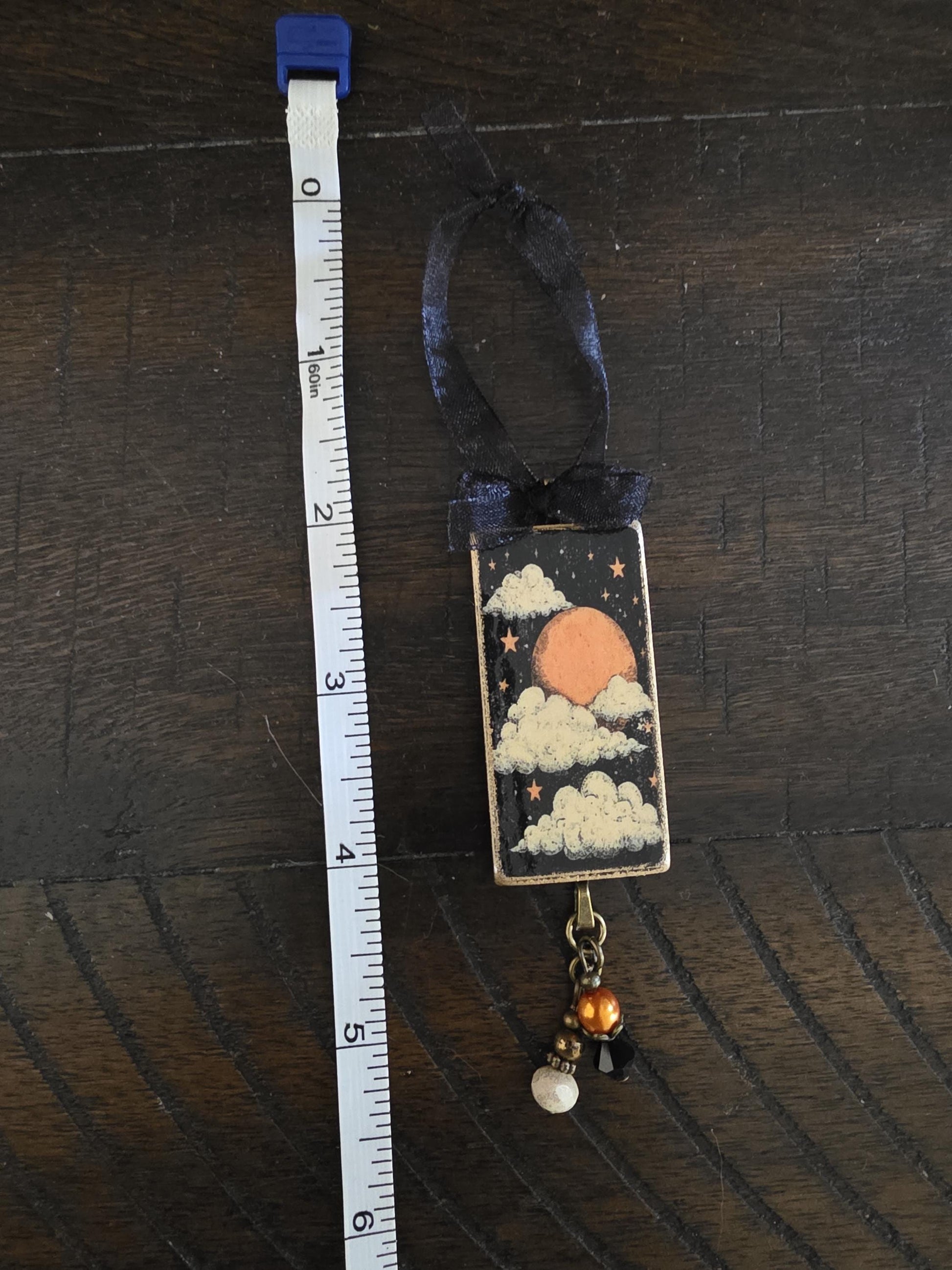 A decorative piece with a black ribbon and a small, colorful design depicting a night sky with a moon and clouds. The piece is placed next to a ruler on a wooden surface.