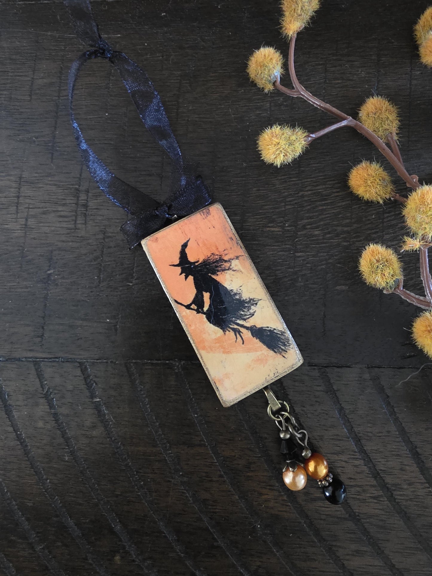 A rectangular pendant with a black silhouette of a witch on a broomstick, hanging from a blue ribbon, and a small black charm with gold beads.