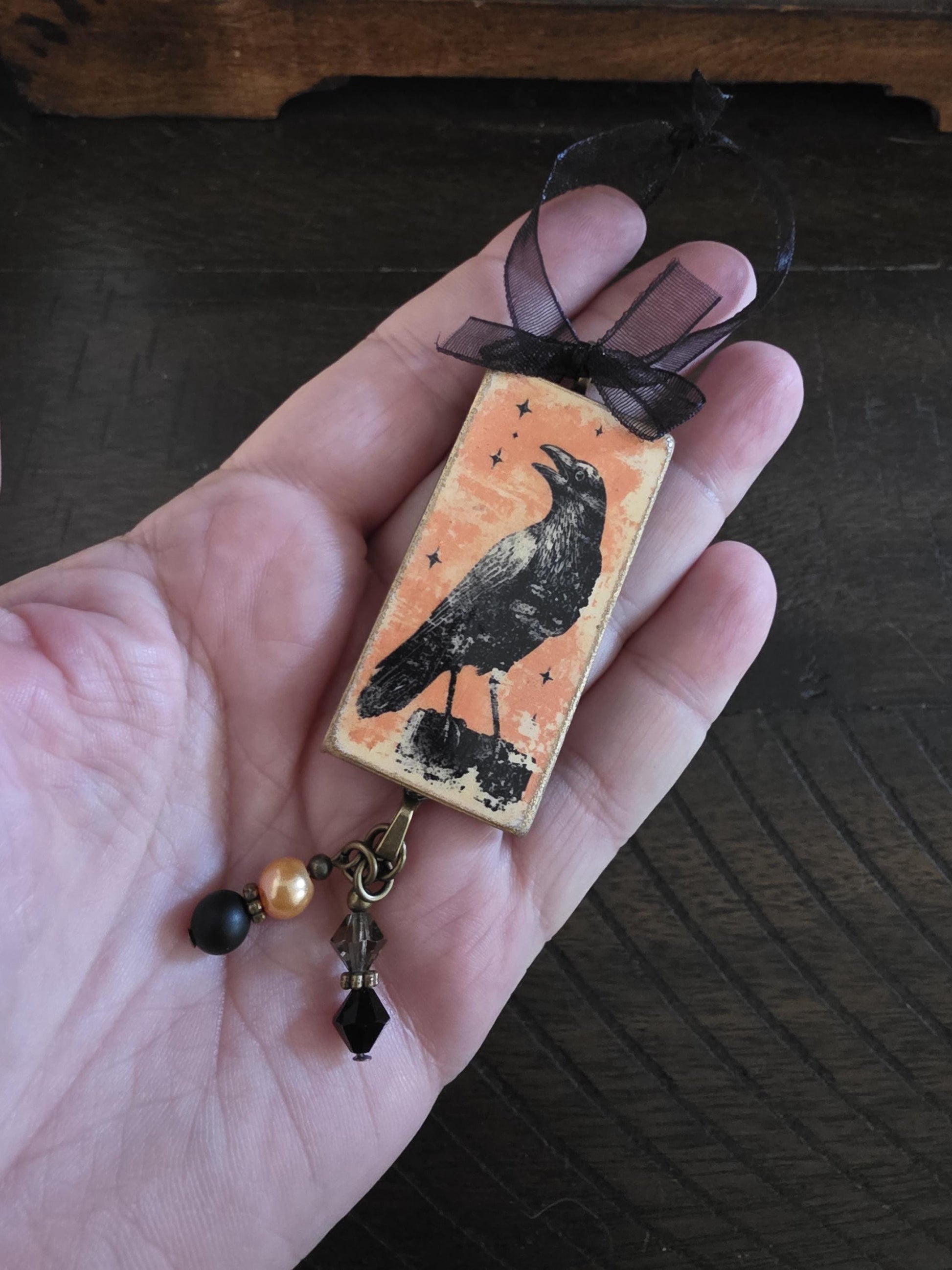 A hand holding a small, rectangular ornament with a black bird image and a black ribbon.