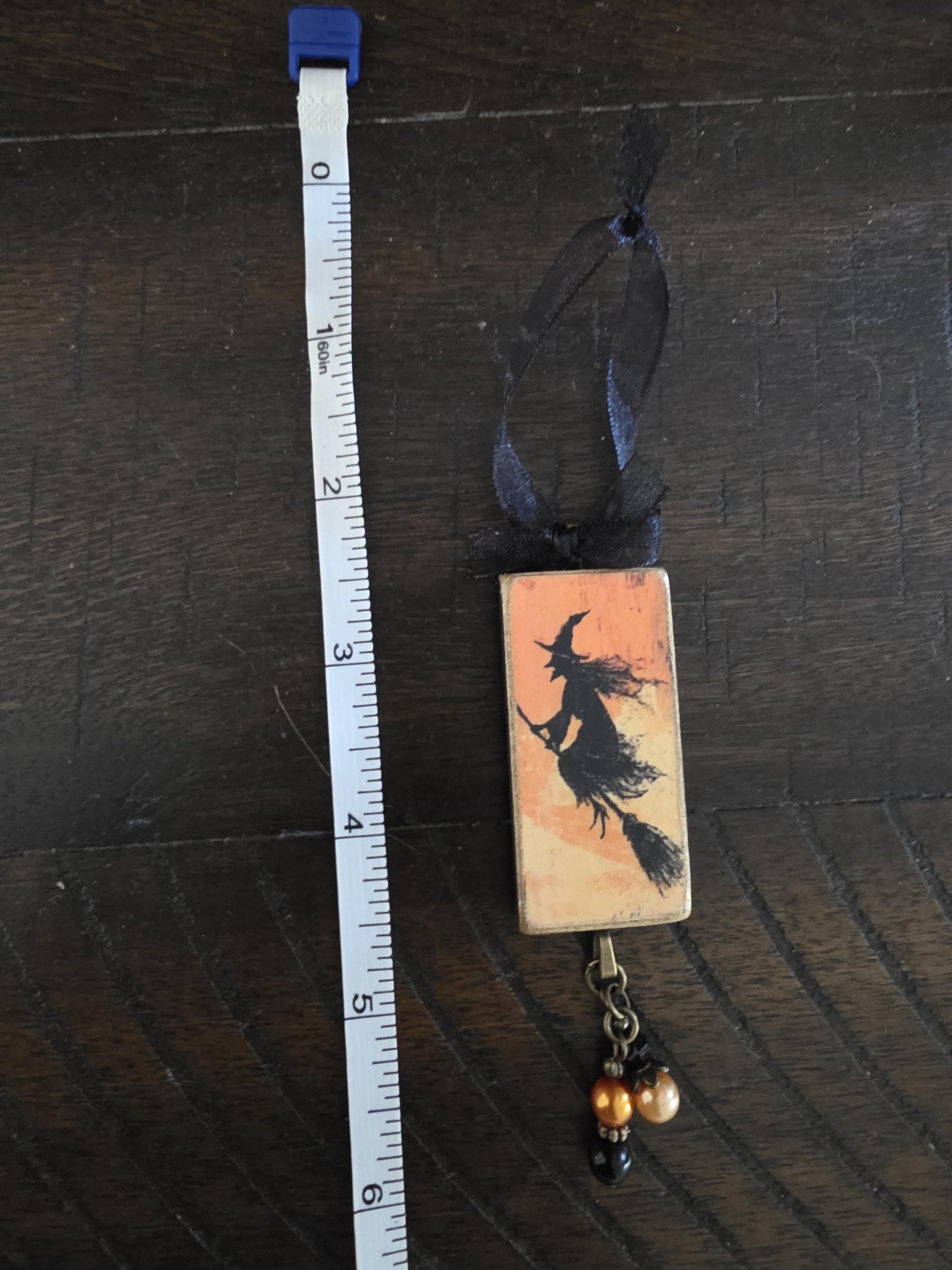 A ruler is placed next to a rectangular charm or ornament that features a silhouette of a witch on a broomstick.