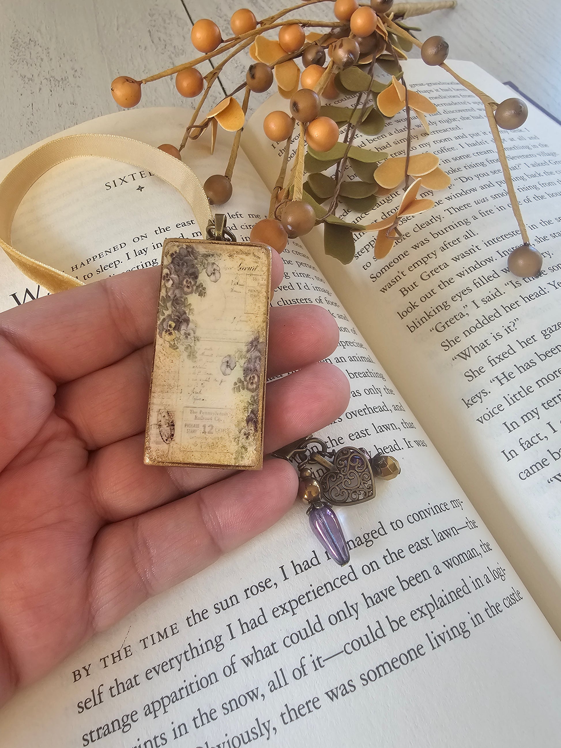 A hand holding a small, rectangular, antique-looking object with a floral design, placed on top of an open book with text visible.