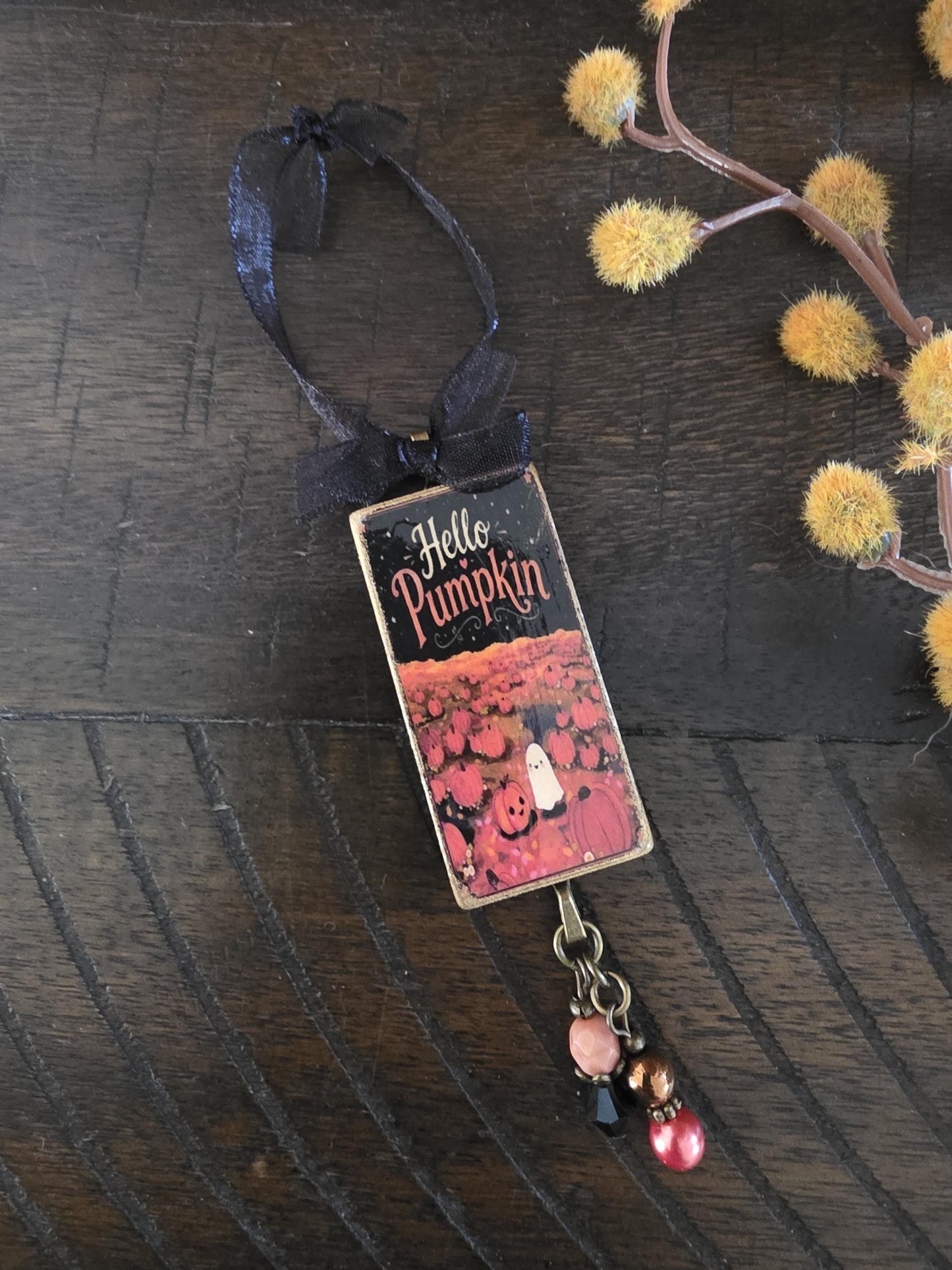 A metallic pendant with a pumpkin and ghost design, featuring the text "Hello Pumpkin" on it, is hanging on a wooden surface.