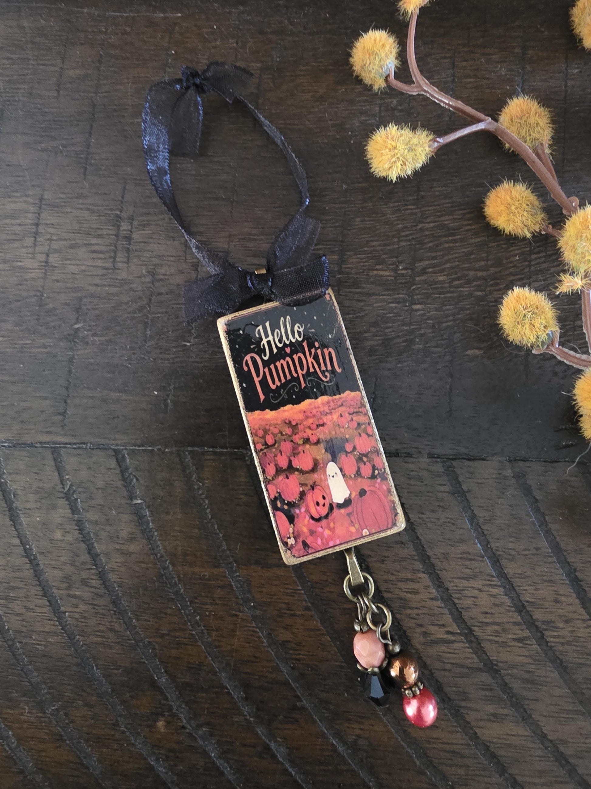 A metallic pendant with a pumpkin and ghost design, featuring the text "Hello Pumpkin" on it, is hanging on a wooden surface.