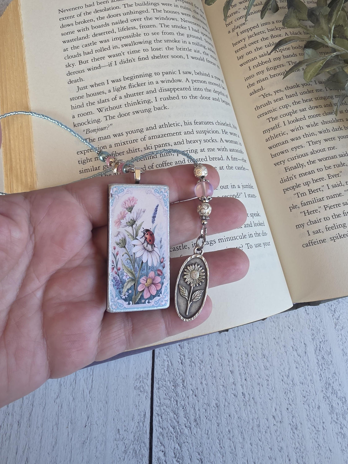 A hand holding a small, colorful book charm with a flower design, resting on an open book with text.
