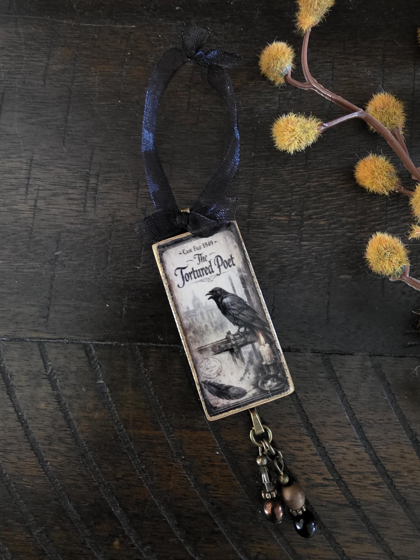 a rectangular pendant with a black and white illustration of a crow perched on a bridge, and a black ribbon hanging from the pendant.