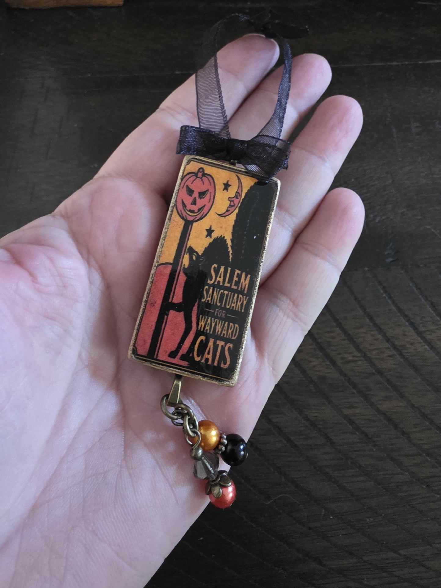A hand holding a small decorative item with a Halloween-themed design, including a jack-o-lantern and a silhouette of a cat.