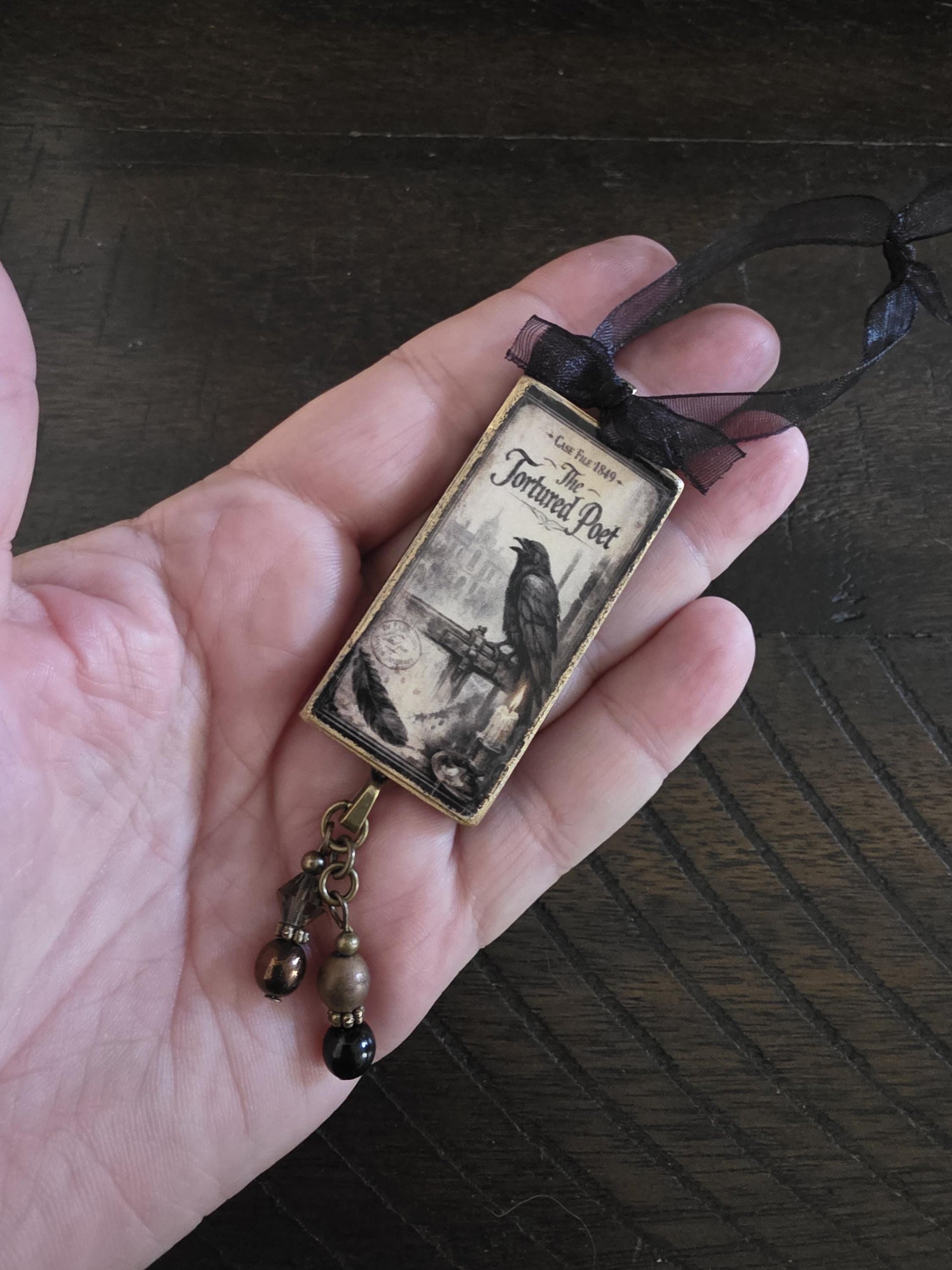 A hand holding a small rectangular pendant with a black ribbon and a picture of a bird, possibly a caged bird, on it.