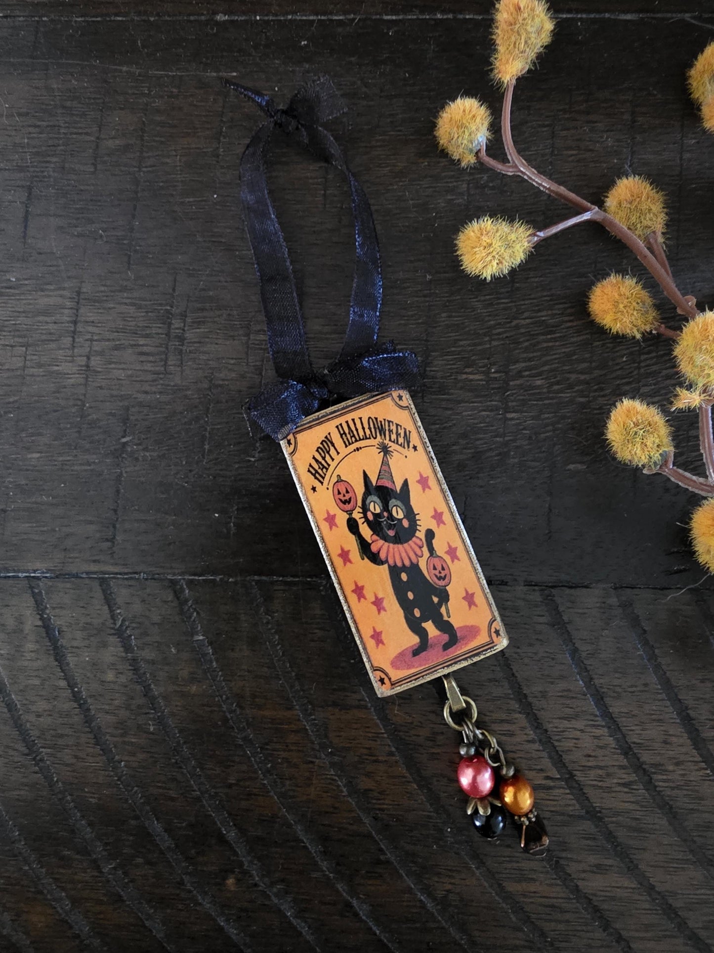A Halloween-themed ornament featuring a black cat wearing a party hat and holding a jack-o-lantern, with a ribbon and beads hanging from it, placed on a wooden surface next to some yellow flowers.