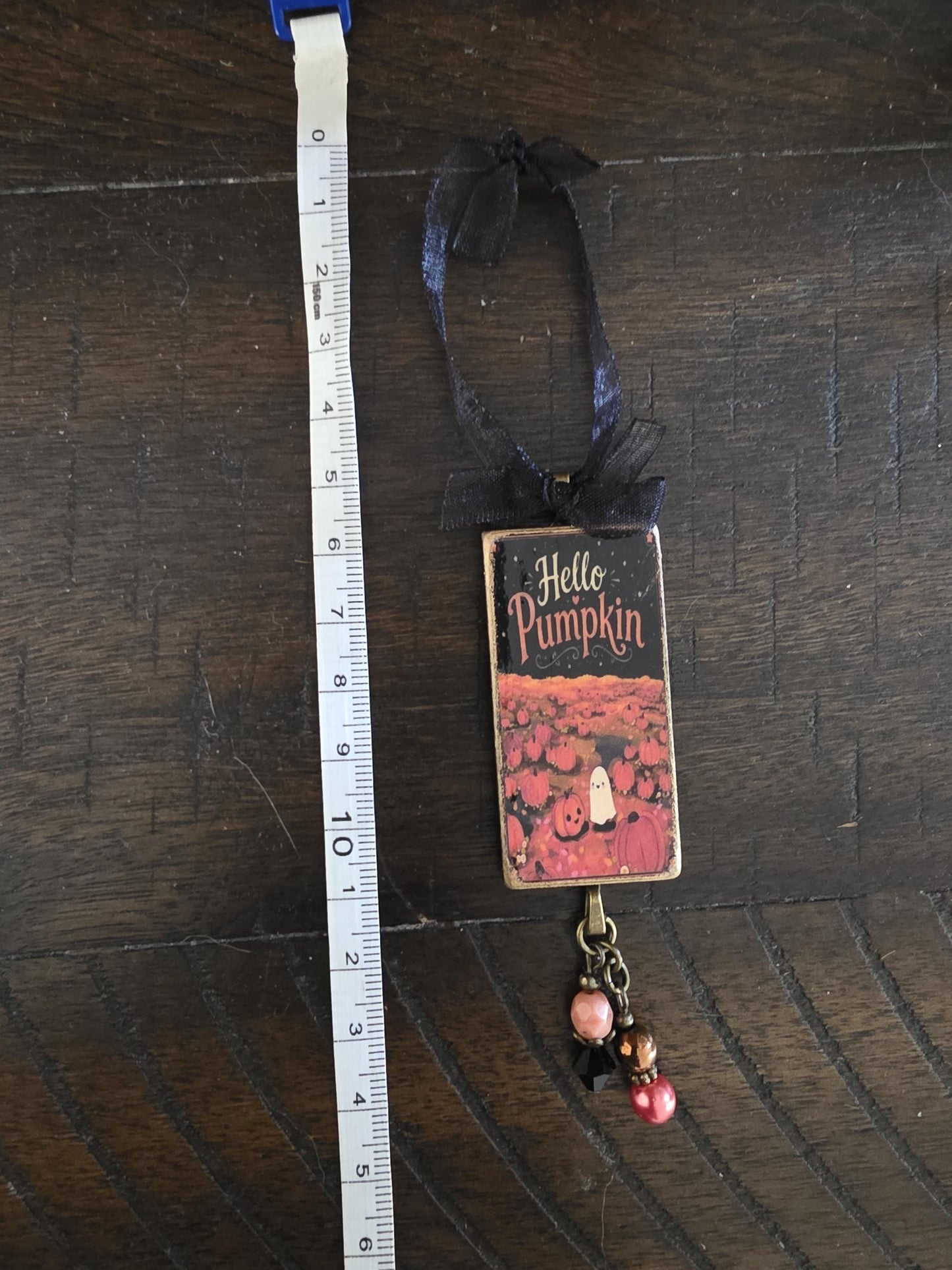 A small decorative item with a pumpkin and ghost design, hanging from a ribbon and measuring its size with a ruler.