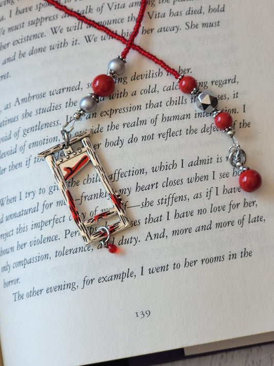 Strung beaded bookmark atop an open book. Bookmark has red and silver glass beads and a silver plated guillotine charm with red enamel paint on the edges and a tiny red glass drop bead.